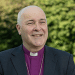 Man outside in a garden, he is wearing a clerical collar wearing a purple shirt and black jacket.