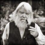 Black and white photo of a man smoking a pipe, he has a white beard and white hair, he's wearing a black waistcoat