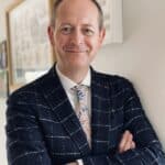 Man wearing a navy checked blazer, shirt and tie, with folded arms, smiling at the camera