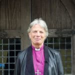 Person in leather jacket with purple clerical shirt, collar and wearing a large cross round their neck