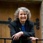 Woman with curly hair, wearing a clerical collar and blue shirt under a grey blazer leans over iron railings in front of what looks like a church door