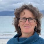 Woman with glasses and a teal jumper on with the sea and mountains in the background
