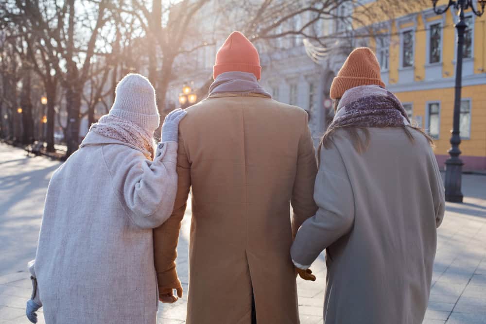 Three people walking away from the camera, they are wearing coats and hats, it looks like winter. They look elderly and are supporting each other.